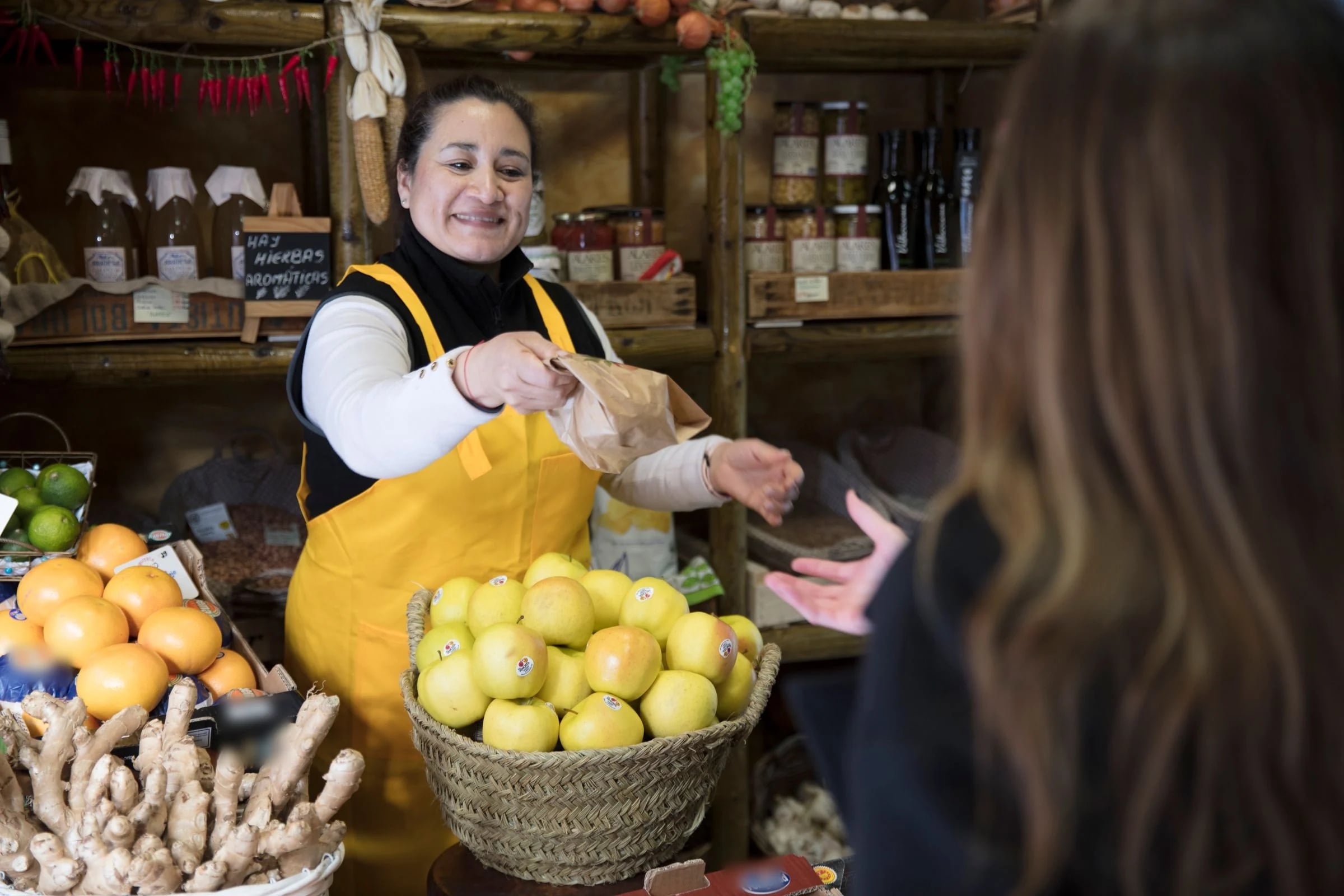Frutera con delantal amarillo entrega una bolsa a una clienta en una tienda de ultramarinos