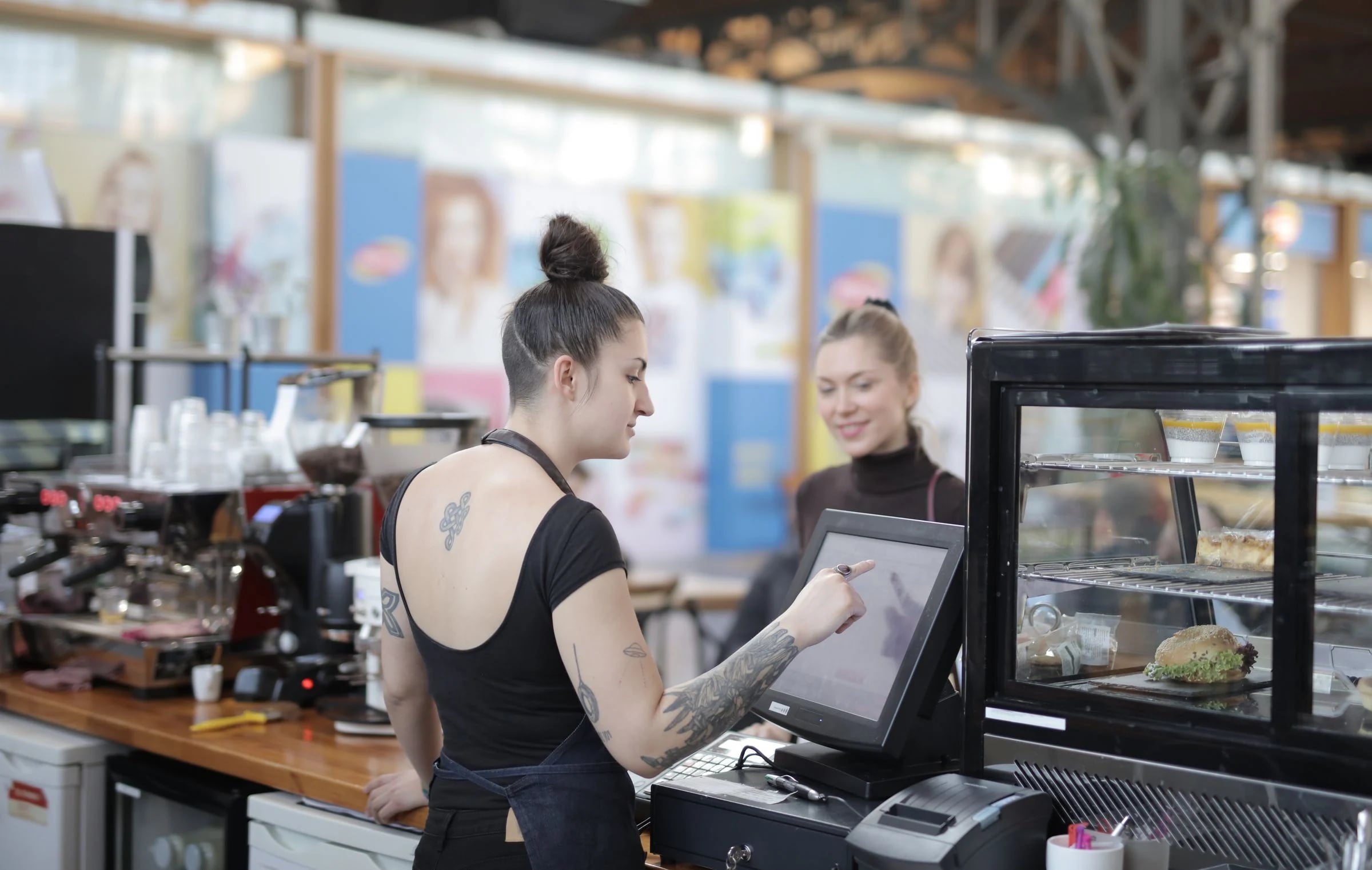 Camarera marca un pedido en la pantalla táctil de la caja de una cafetería, con la cafetera al fondo