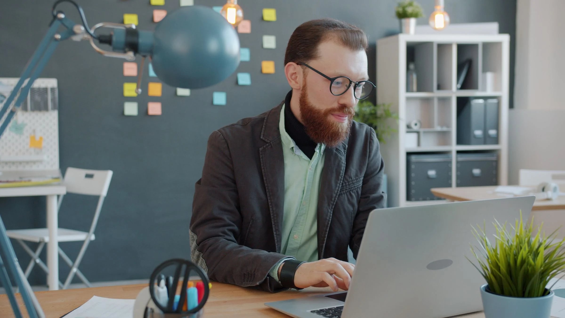 Profesional con gafas y barba trabaja con el portátil en una oficina luminosa con estanterías y plantas