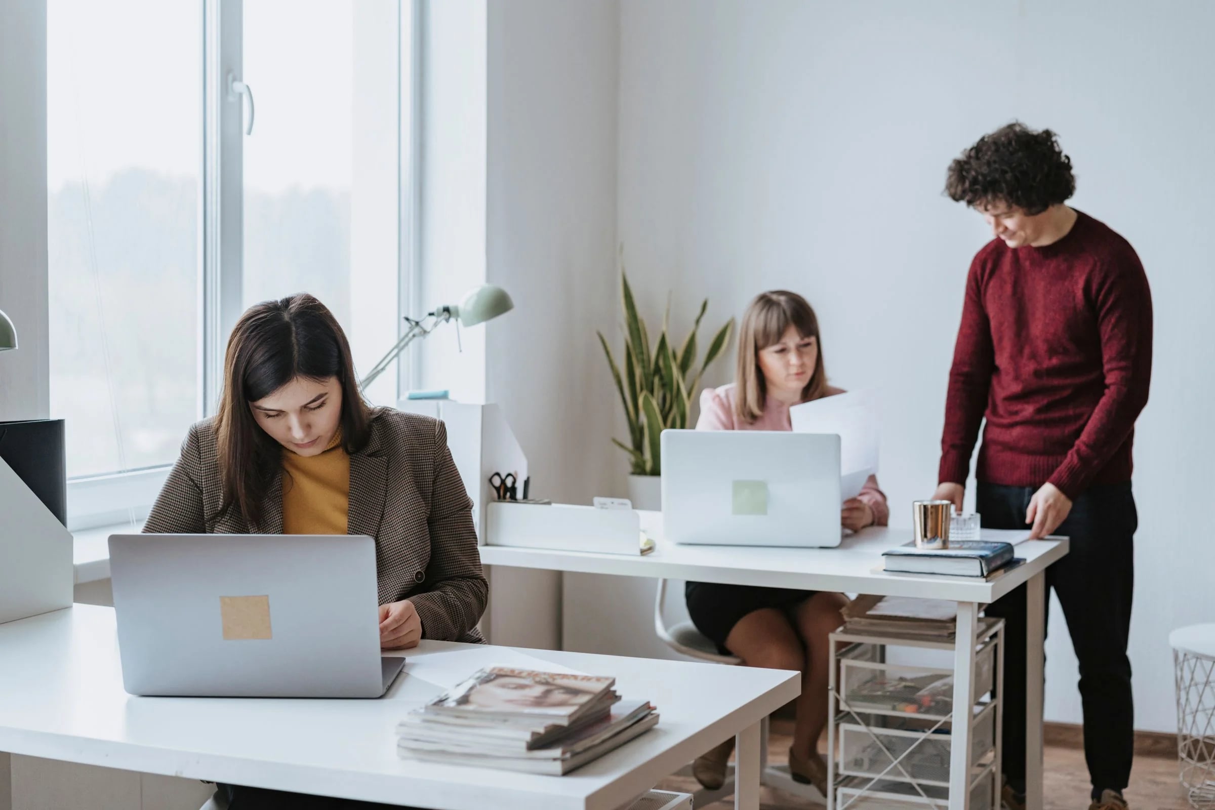 Tres personas trabajando con portátiles en una oficina blanca y luminosa junto a un ventanal