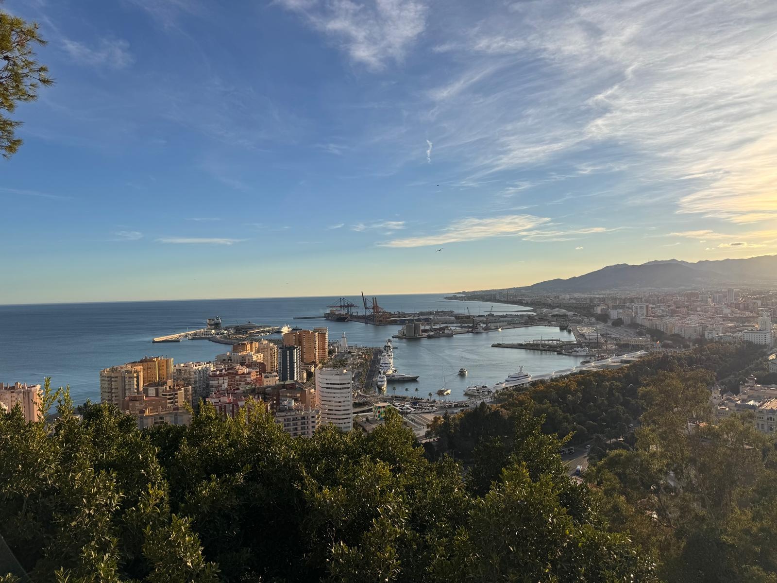 El puerto de Málaga, La Farola y el centro de la ciudad vistos desde Gibralfaro, con los montes al fondo bajo un cielo azul