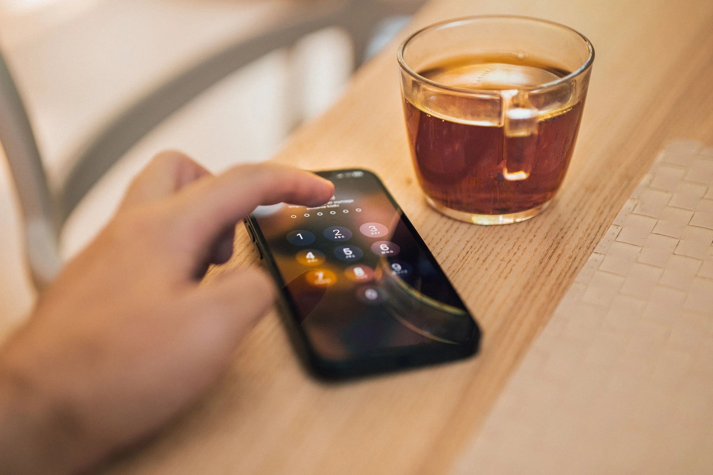 Mano introduciendo el código de desbloqueo en un móvil sobre una mesa de madera junto a un vaso de té