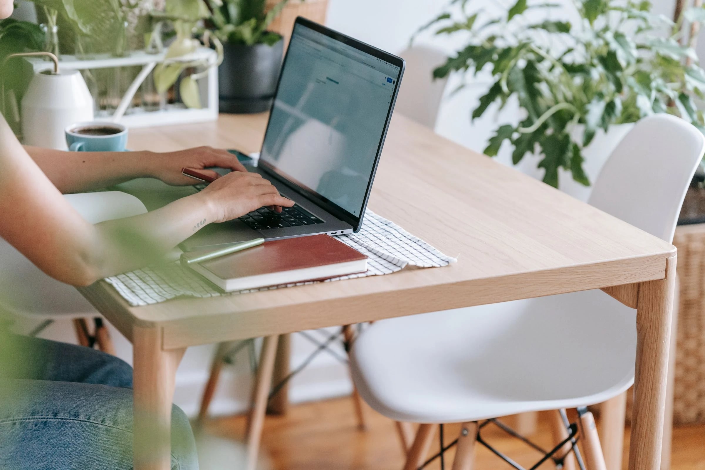 Persona escribiendo en un portátil sobre una mesa de madera con plantas, café y libreta junto a la ventana